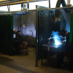 Dwaj spawacze pracują w oddzielnych stanowiskach. Jeden z nich wykonuje spawanie stali nierdzewnej ze zwykłą, tworząc jasny łuk spawalniczy.