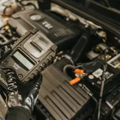 Mechanik w rękawiczkach sprawdza stan akumulatora za pomocą testera. To kluczowy etap diagnostyki, zapobiegający problemom z rozruchem, czyli gazowanie akumulatora.