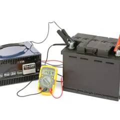 Prostownik ładuje akumulator samochodowy. Obok stoi żółty multimetr pokazujący napięcie 14.64V.