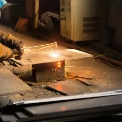 Ręce w rękawicach trzymają palnik, pokazując, jak spawać metal. Iskry lecą podczas pracy.