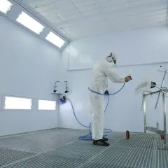 Pracownik w kombinezonie lakierniczym maluje element karoserii w nowoczesnej kabinie lakierniczej.