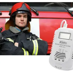 Strażak w hełmie i masce obok czujnika gazu Xblitz. Urządzenie może włączyć alarm bez powodu, sygnalizując potencjalne zagrożenie.