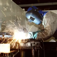 Spawacz w masce i fartuchu pracuje przy metalowych elementach, osiągając temperaturę topnienia stali czarnej. Iskry sypią się wokół.