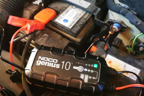 Ładowarka NOCO Genius 10 podłączona do akumulatora samochodowego. Zielona lampka sygnalizuje postęp ładowania, rozwiązując problem zbyt długiego ładowania akumulatora.