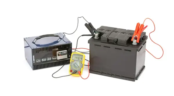 Prostownik ładuje akumulator samochodowy. Obok stoi żółty multimetr pokazujący napięcie 14.64V.