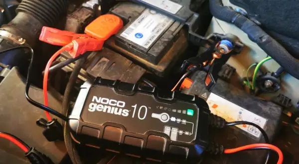 Ładowarka NOCO Genius 10 podłączona do akumulatora samochodowego. Zielona lampka sygnalizuje postęp ładowania, rozwiązując problem zbyt długiego ładowania akumulatora.