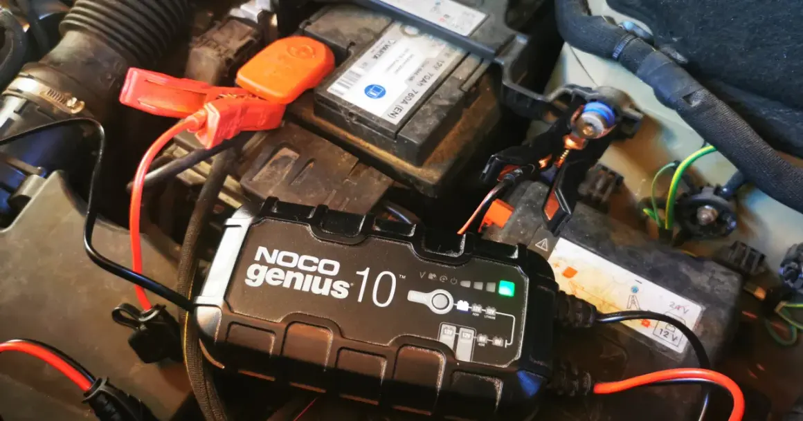 Ładowarka NOCO Genius 10 podłączona do akumulatora samochodowego. Zielona lampka sygnalizuje postęp ładowania, rozwiązując problem zbyt długiego ładowania akumulatora.