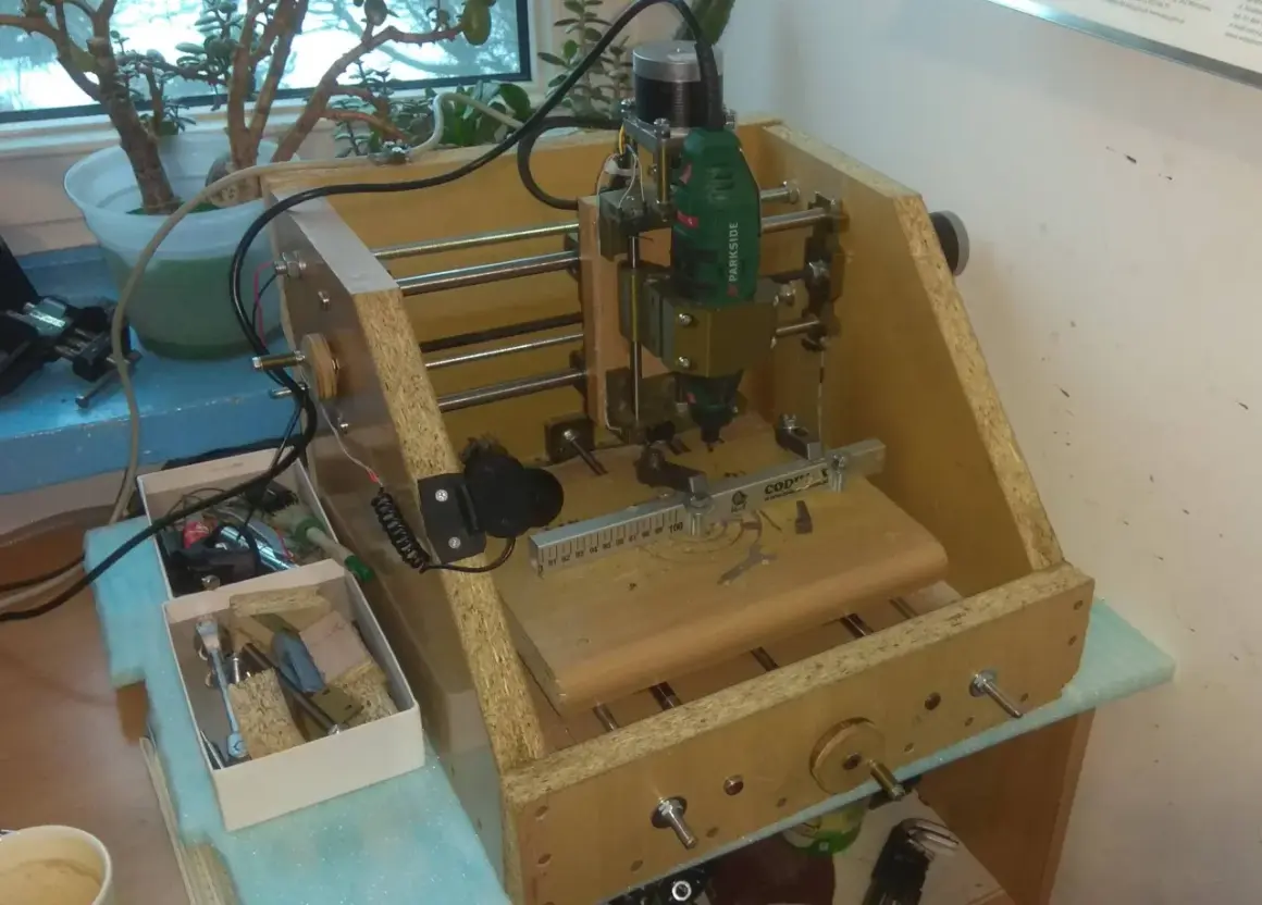 Domowa budowa frezarki CNC z narzędziami i rośliną w tle.