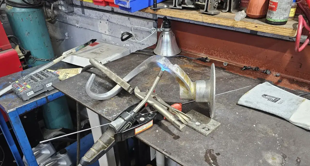 Demonstracja jak spawać aluminium: palnik z niebieskim płomieniem ogrzewa metalowy element, obok szczypce i drut spawalniczy.