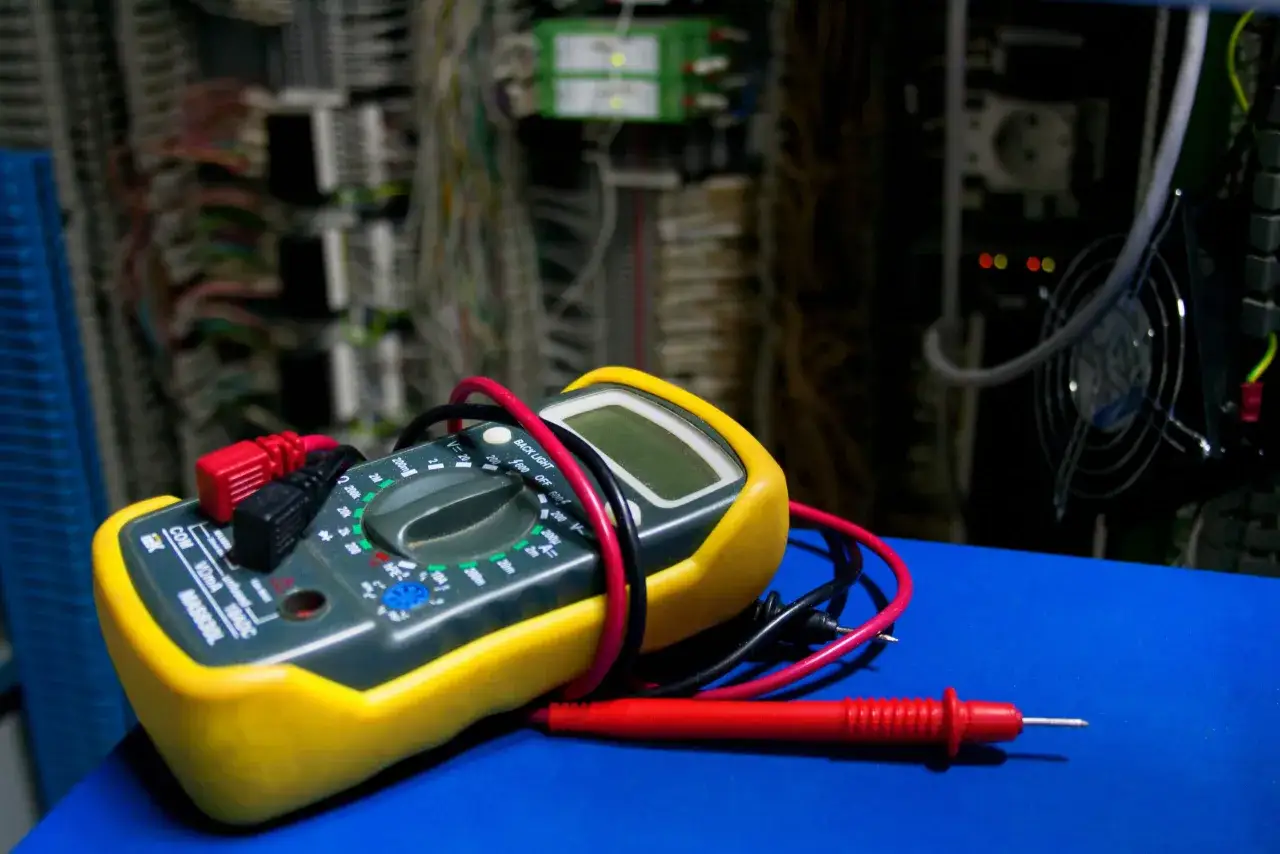 Ż&oacute;łty multimetr z czerwonym i czarnym przewodem leży na niebieskiej powierzchni. W tle widać szafę elektryczną, kt&oacute;ra pomoże Ci dowiedzieć się, jak obliczyć napięcie.